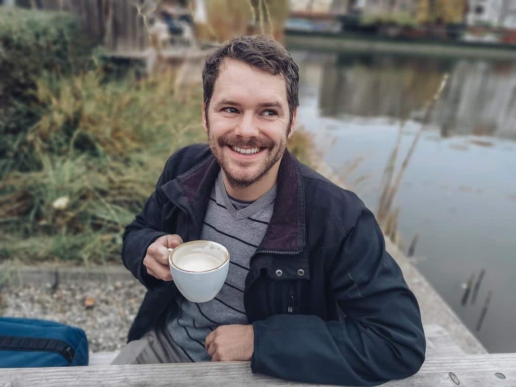 Man with full head of hair outside drinking coffee and smiling